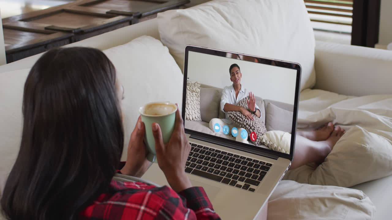 African american woman holding a coffee cup having a video call on laptop sitting on couch at home