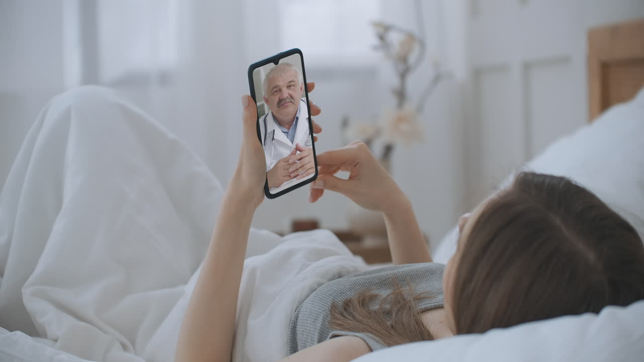 Young Girl lie in bed at Home Using Smartphone to Talk to Her Doctor via Video Conference Medical App. Beautiful Woman Checks Possible Symptoms with Professional Physician Using Online Video Chat.