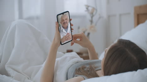 молодая девушка лежит в постели дома, используя смартфон, чтобы поговорить со своим врачом через видеоконференцию медицинское приложение. красивая женщина проверяет возможные симптомы с профессиональным врачом, используя онлайн видеочат.