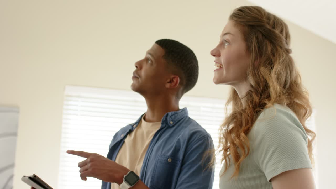 Diverse couple tapping, swiping tablet with smartwatch in living room noticing ceiling and pointing