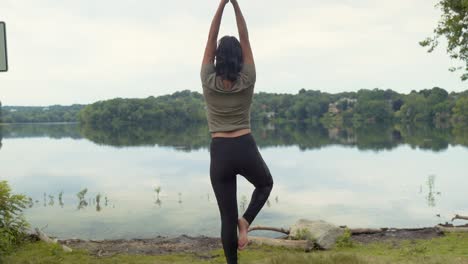 latina woman relaxed practicing yoga in tree pose meditating on rock by pond