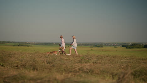 dos mujeres con un perro caminando a través de vastas tierras de cultivo, una sosteniendo la correa del perro mientras el perro trata de correr adelante, acompañadas por una madre e hija caminando cerca en el campo abierto