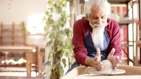 focused senior biracial potter with long beard using potter's wheel in pottery studio, slow motion