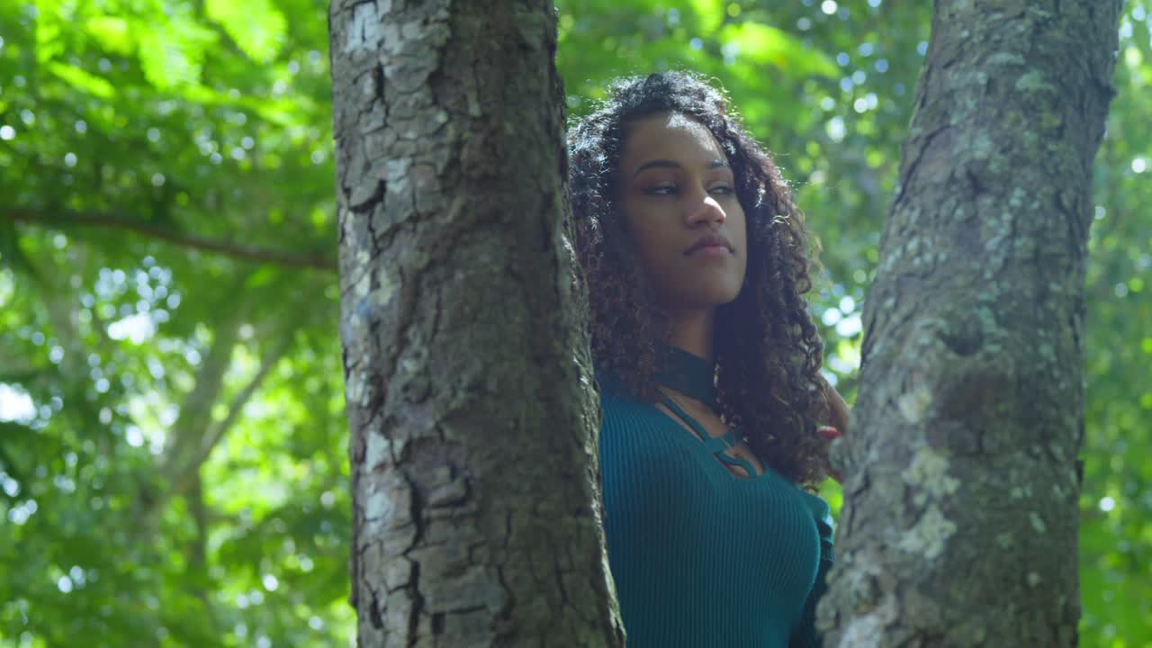 una joven de cabello rizado con la luz del sol brillando en su cabello y cara mientras está de pie detrás de un tronco de árbol