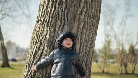 un niño joven está de pie con la espalda apoyado en un gran tronco de árbol, mirando hacia arriba con una expresión curiosa, sus brazos están ligeramente extendidos, y está vestido con una chaqueta negra brillante