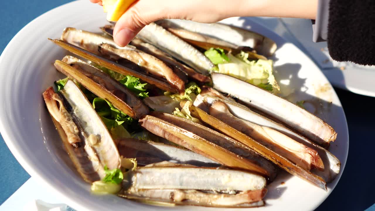 Käsi asettamassa puristettua sitruunaa grillattujen simpukoiden päälle valkoisella lautasella salaatin kera
