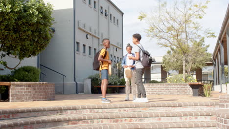 multiracial students with backpacks chatting happily outside school building on sunny day