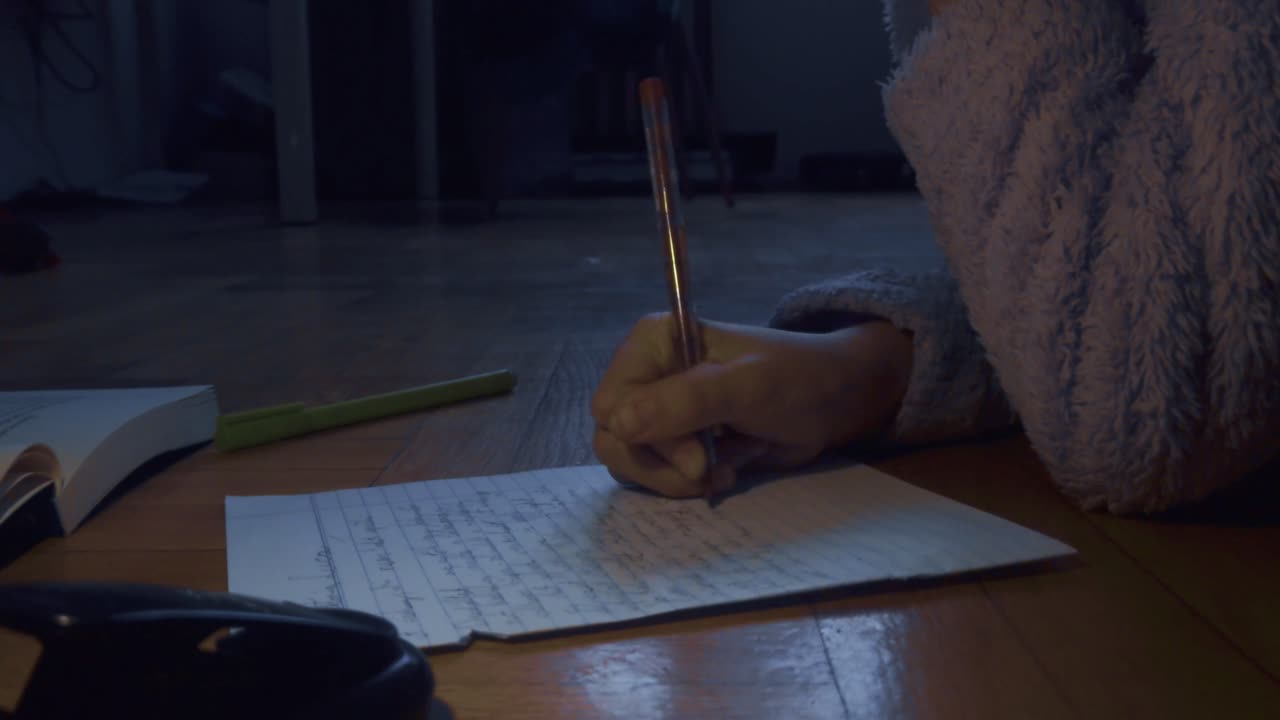 In a dimly lit room, a hand captures thoughts on paper with a pen. The scene features a cozy blue robe and wooden flooring, embodying the quiet essence of late-night reflection and creative focus