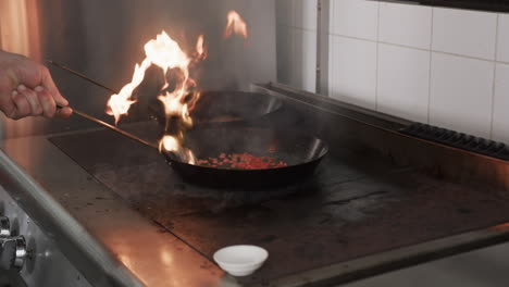 caucasian male chef frying food in frying pan with bursting fire in kitchen, slow motion