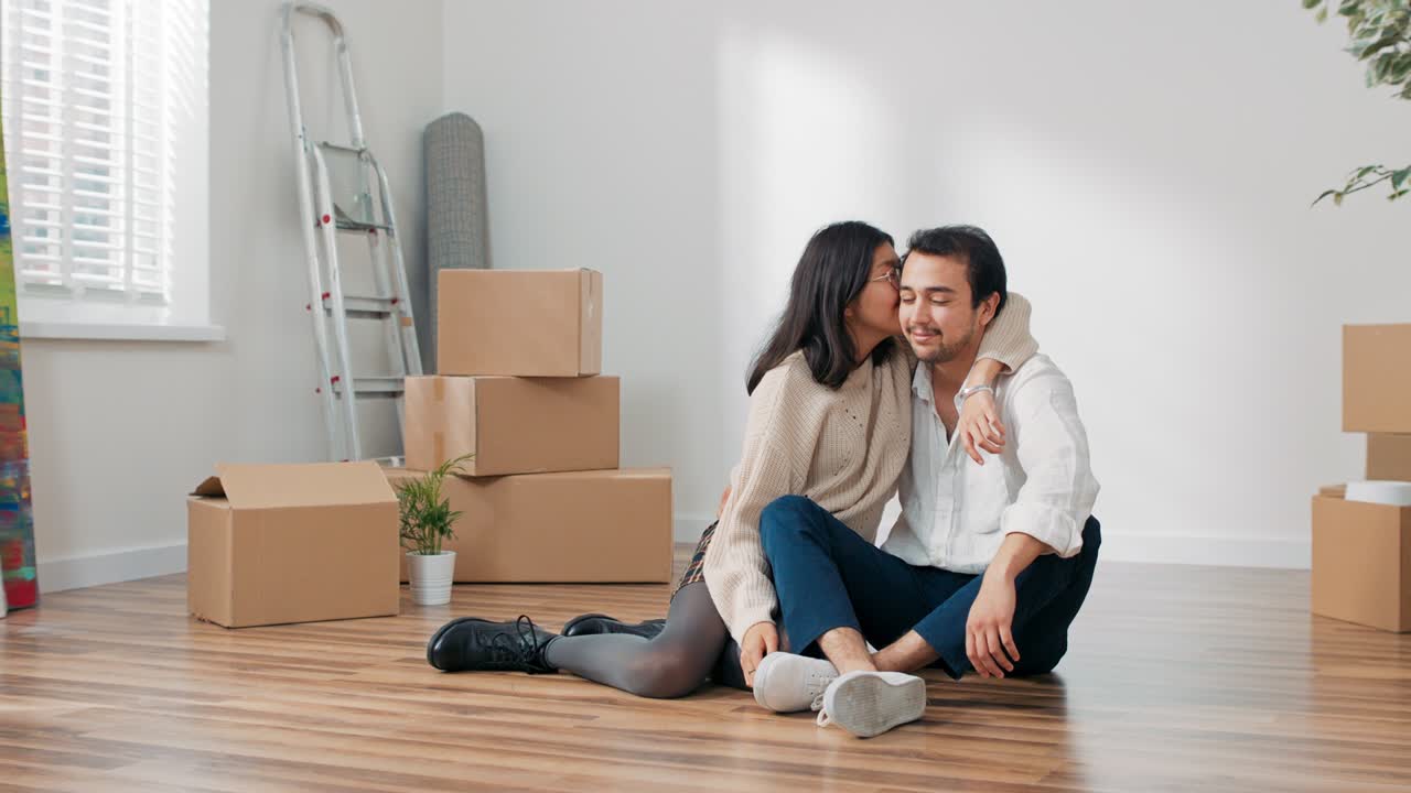 Happy contented young couple moves into their first apartment together, couple sits on a wooden floor close together, an empty room, boxes of unpacked stuff strewn everywhere, freshly renovated
