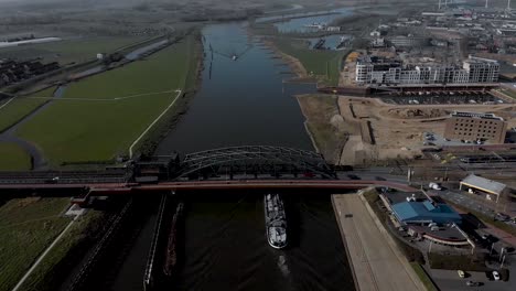 Aerial-descend-showing-a-large-cargo-ship-passing-beneath-the-iron-draw-bridge-revealing-the-cityscape-quay-of-Hanseatic-medieval-city-of-Zutphen,-the-Netherlands-along-the-river-IJssel