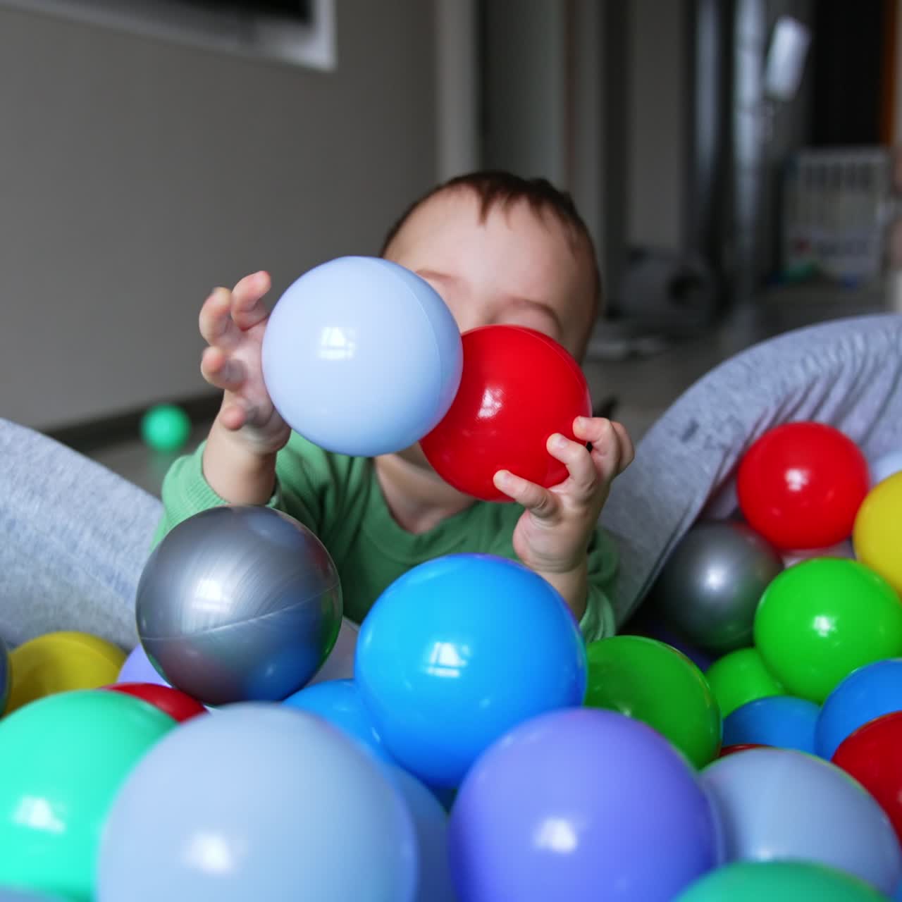 Playful Caucasian kid banging bright balls from a dry basin. Little baby having good time at home. Blurred backdrop