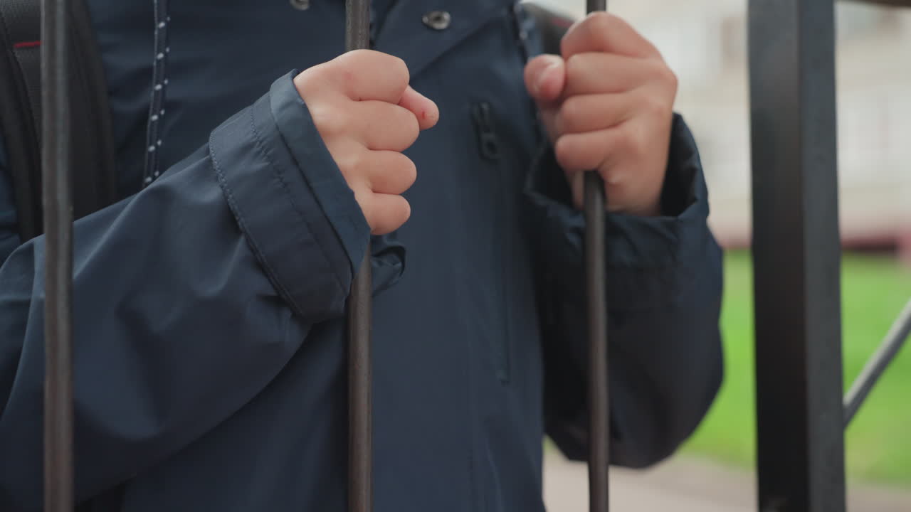 Okul kapısında çantasını hazırlayan genç çocuk. Metal parmaklıkları kavrayan ellerin yakın çekimi, lacivert ceket ve kapüşon, arka planda banliyö okul binası, sabah rutini, tereddütlü ifade, sessiz beklenti
