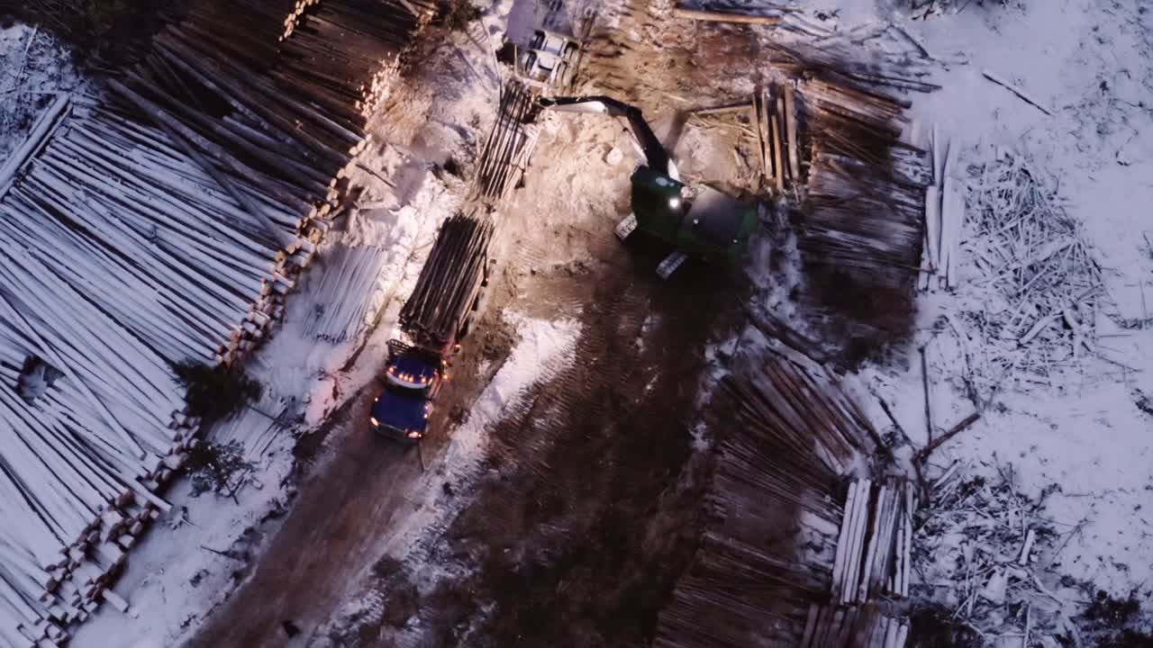 Aerial top down drone cranes down as timber manipulator fills up timber transportation truck from nearby snow covered cut down mature pine trees piles