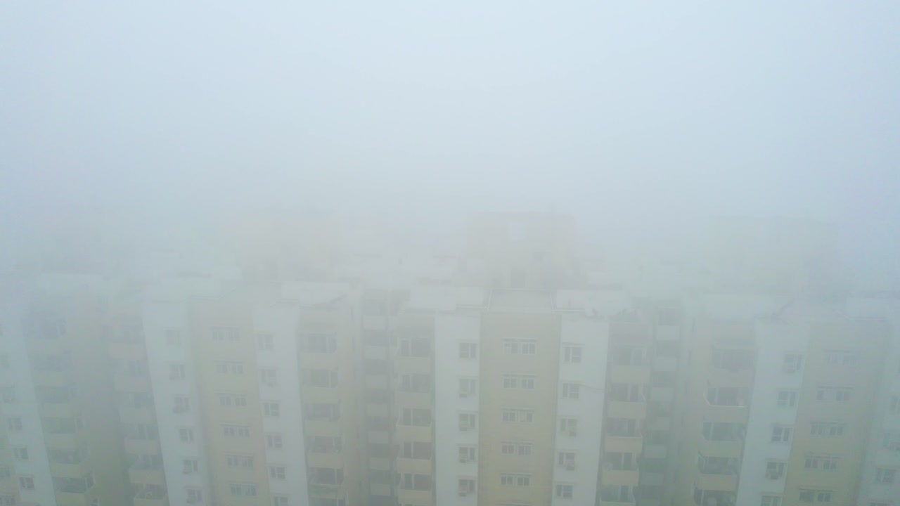 Aerial drone shot flying back from brown white skyscraper as dense fog sheets roll around it in a surreal scene in Delhi India