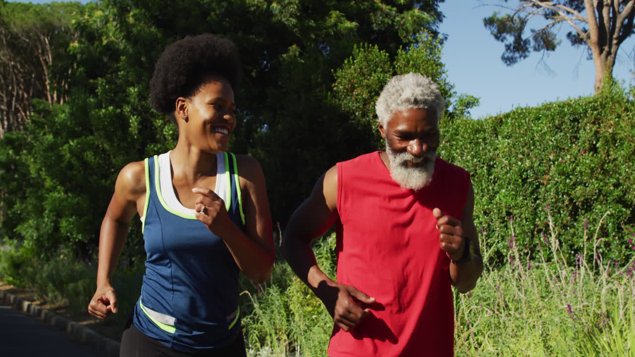 una pareja de ancianos afroamericanos sonrientes haciendo ejercicio al aire libre corriendo en una carretera verde soleada