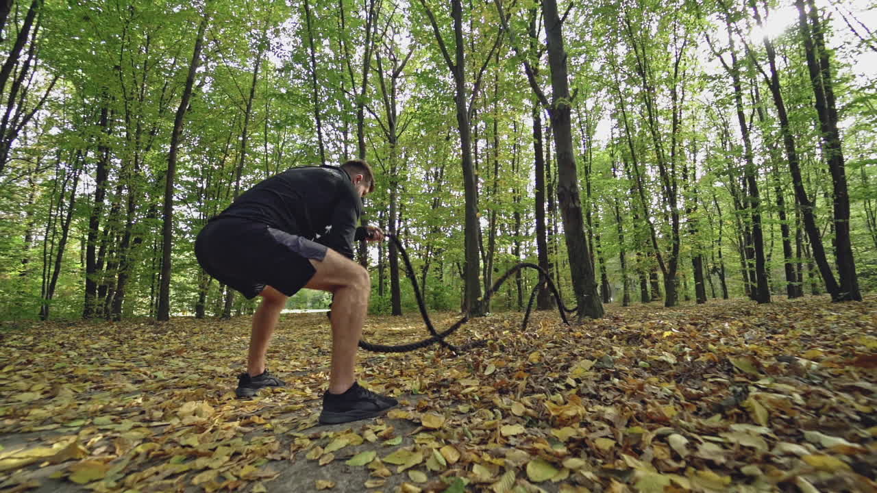 Sportsman doing workout with a rope outdoors. Muscular athlete exercising with battle ropes on the autumn forest background. High intensive workout.