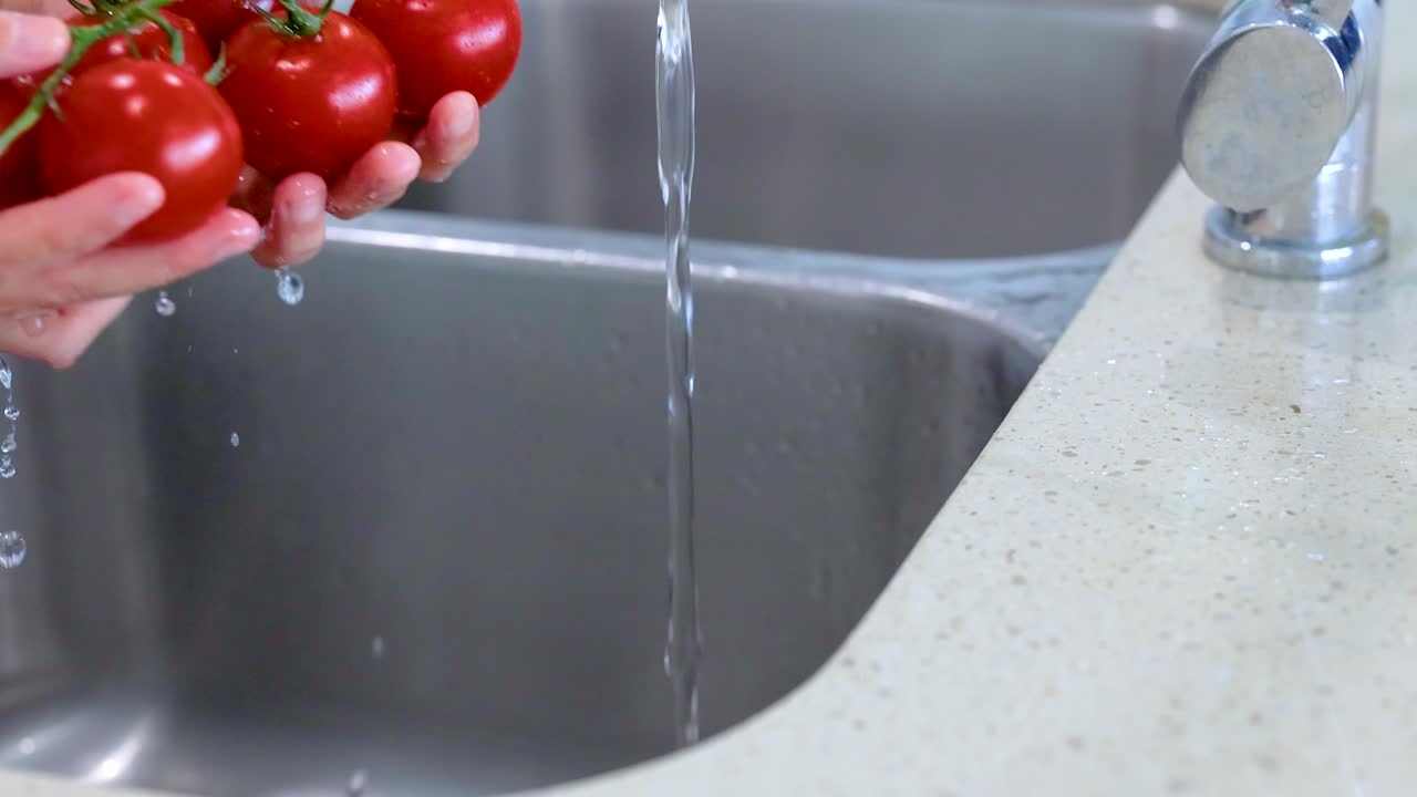 Hands rinse fresh tomatoes under running water in a kitchen sink. Bright lighting enhances the clean, domestic setting