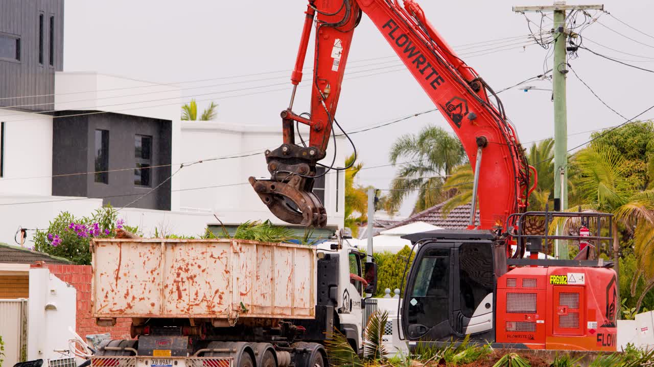 A red hydraulic excavator transfers palm plants and debris into a large skip bin beside modern houses in daylight, with steady camera and overcast lighting