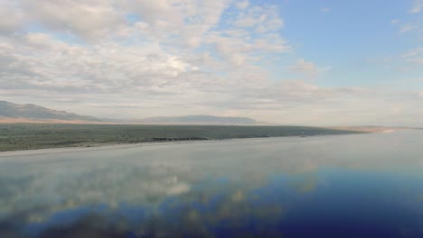 Salt-lake-and-mountains,-salt-flats-reflecting-sun,-clouds-and-blue-sky,-symmetry-reflective-aerial-landscape-panoramic-movement,-sunny-weather,-pretty-cloud,-shot-in-Salton-Sea