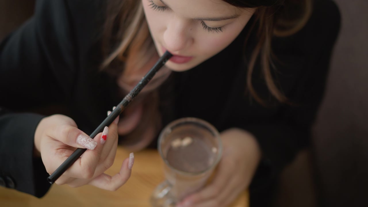 Top-down close-up of seated person bringing patterned straw to mouth while steadying clear glass filled with liquid and ice