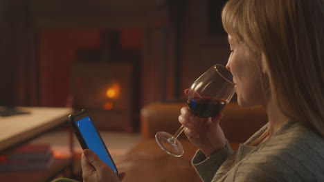 woman at home sitting on sofa with fire with blue screen mobile phone holding glass of wine