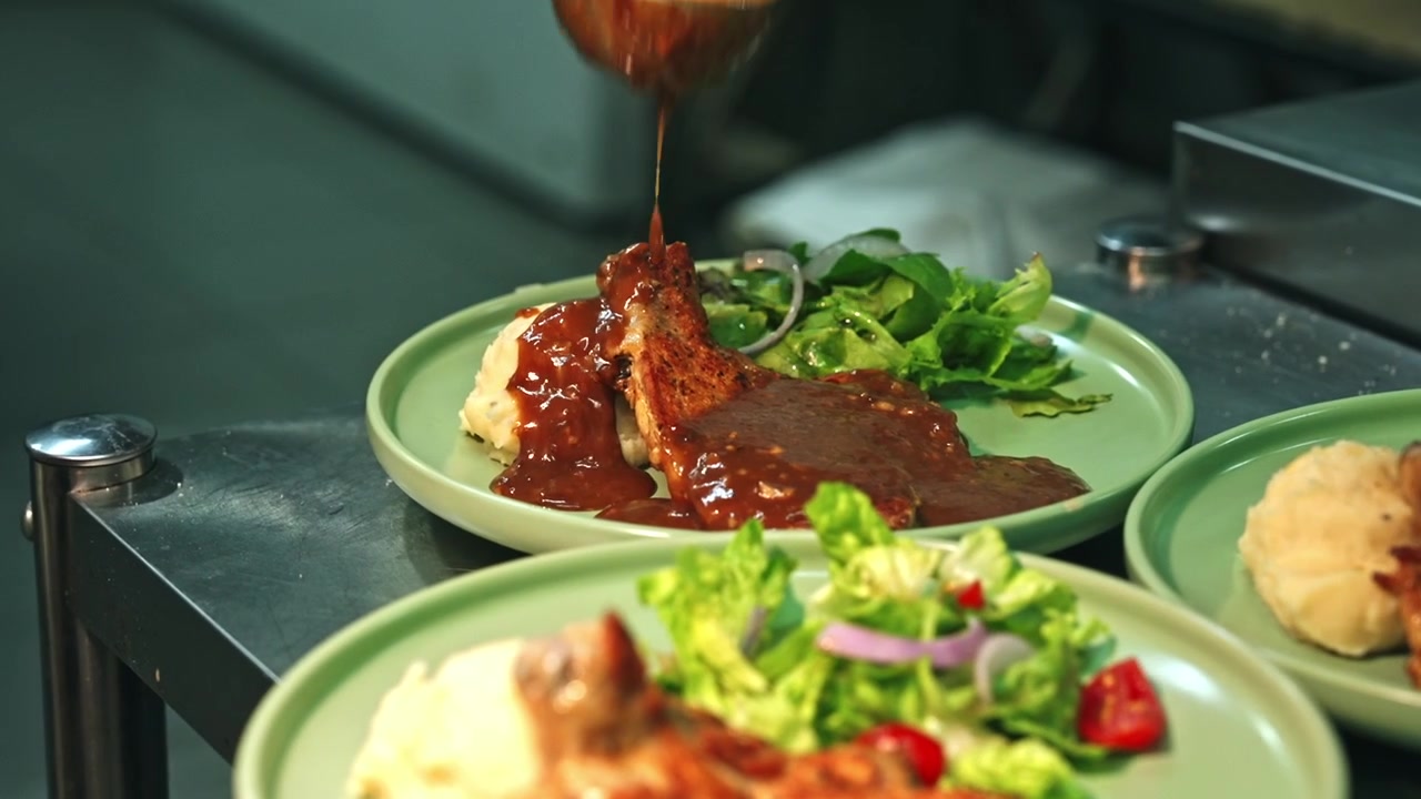 Close-up tracking shot of a chef ladling rich brown sauce over grilled chicken chops served on round green plates with mashed potatoes and green salad.