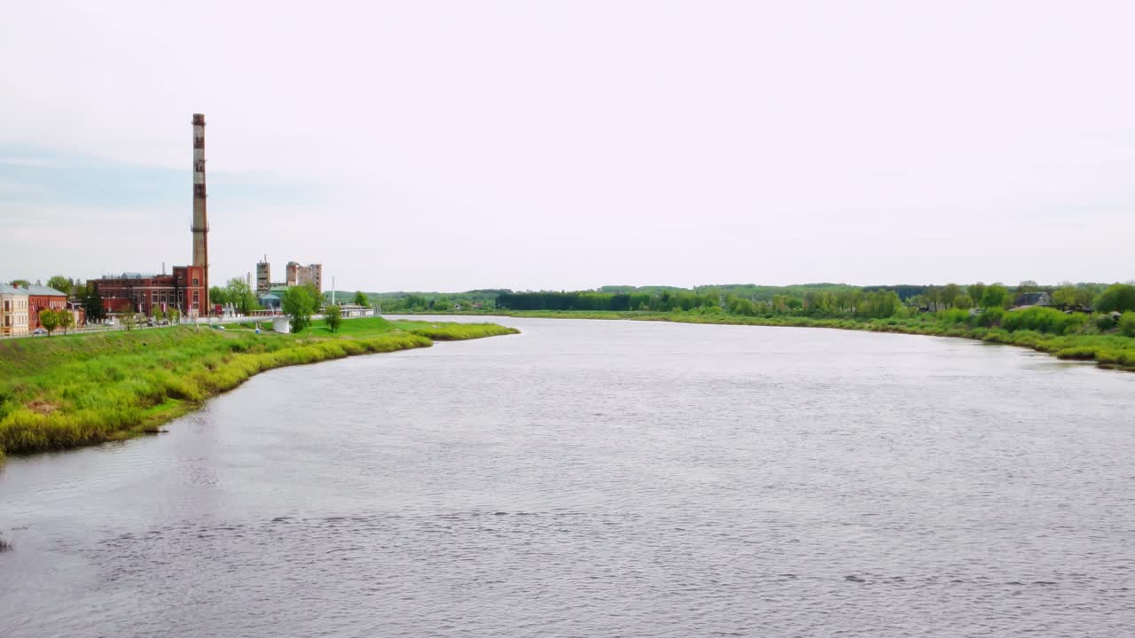 Wide river with grassy banks and a tall factory chimney under a pale sky. Industrial structures on the horizon. Shot in Daugavpils, Latvia, Baltics