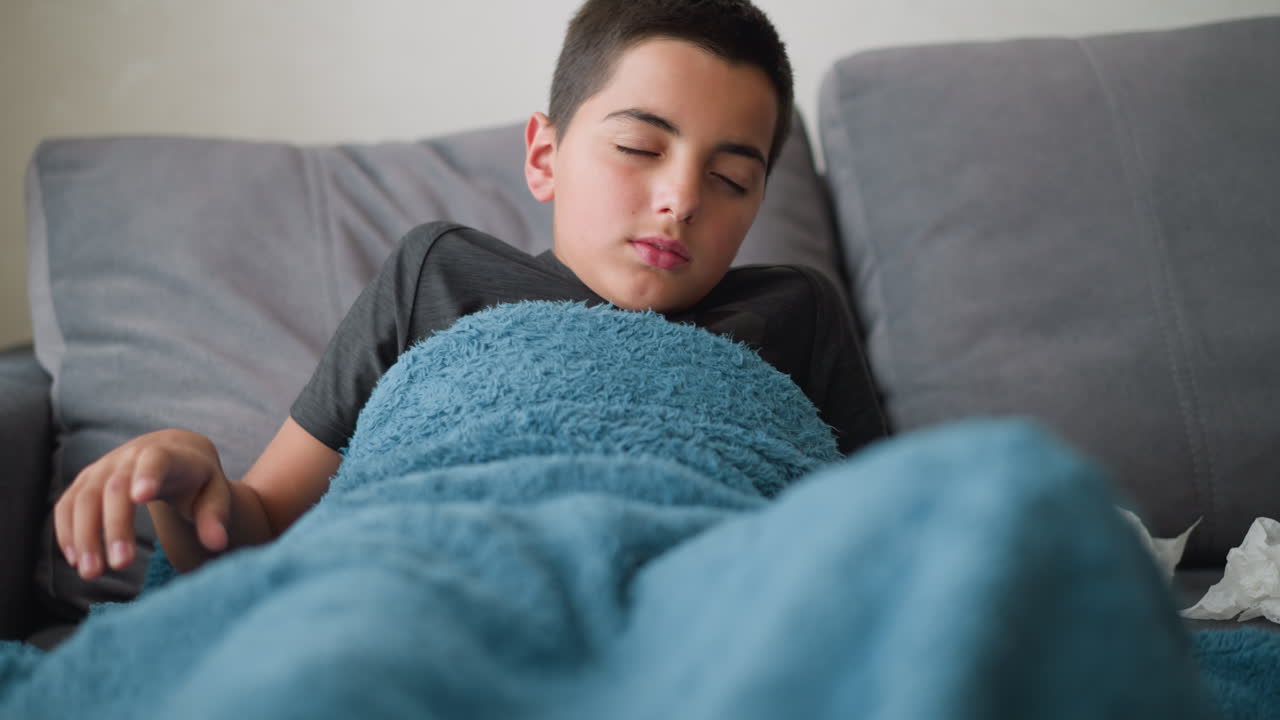 Sick student on holiday resting on couch wrapped in blanket adjusting for comfort with used tissue beside him, relaxing indoor setting with soft natural light streaming through window