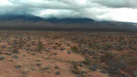 Aerial-shot-flying-low-to-the-ground-of-a-fenced-off-location-filled-with-desert-shrubs-and-the-Rocky-Mountains-in-the-background-with-thick-clouds-overhead