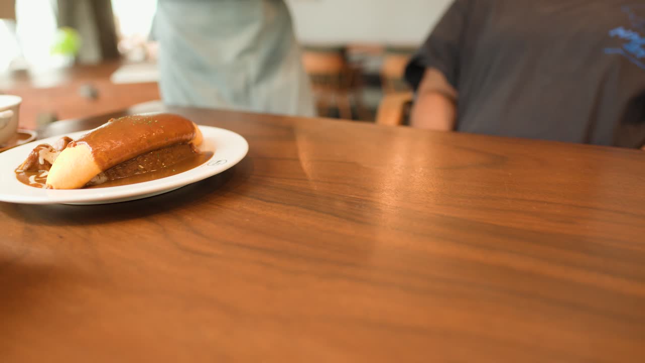 Server sets two omurice curry plates on wooden table in bright, casual restaurant setting