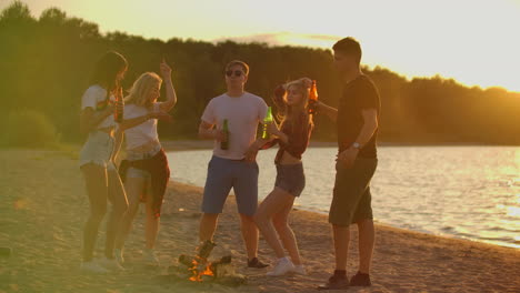 Die-Jungen-Studenten-Verbringen-Den-Sommerabend-Bei-Sonnenuntergang-In-Shorts-Und-T-Shirts-Am-Strand-Am-Lagerfeuer-Mit-Bier.-Sie-Tanzen-Und-Genießen-Die-Aussicht-In-Der-Natur.