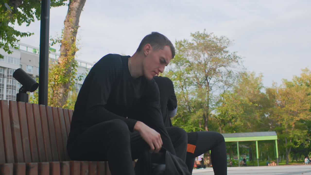 Young man alone in park, Caucasian male seated on city bench, Reflective young man sitting among autumn trees, Casual scene of young man with backpack contemplating in urban park during fall