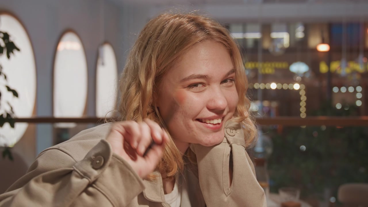 Primer plano de mujer joven aplicándose maquillaje con brocha de delineador con iluminación suave. La mujer lleva una chaqueta beige e interactúa casualmente sosteniendo una pequeña herramienta cerca de su rostro durante toda la escena. Fondo