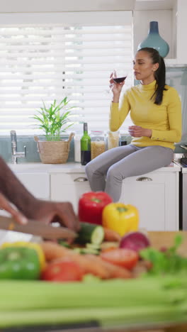 video vertical de una mujer biracial feliz bebiendo vino en la cocina mientras su pareja prepara la comida