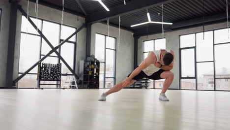 fitness fanatic performing lunge with upper body twists warm up exercise in bright modern gym with white tank and black shorts balancing on light wooden floor beneath hanging straps and large windows