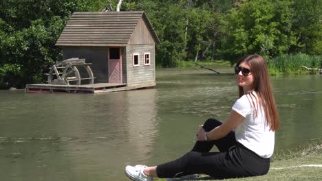 Young-white-girl-sitting-next-to-old-cabin-on-small-river-and-smiling