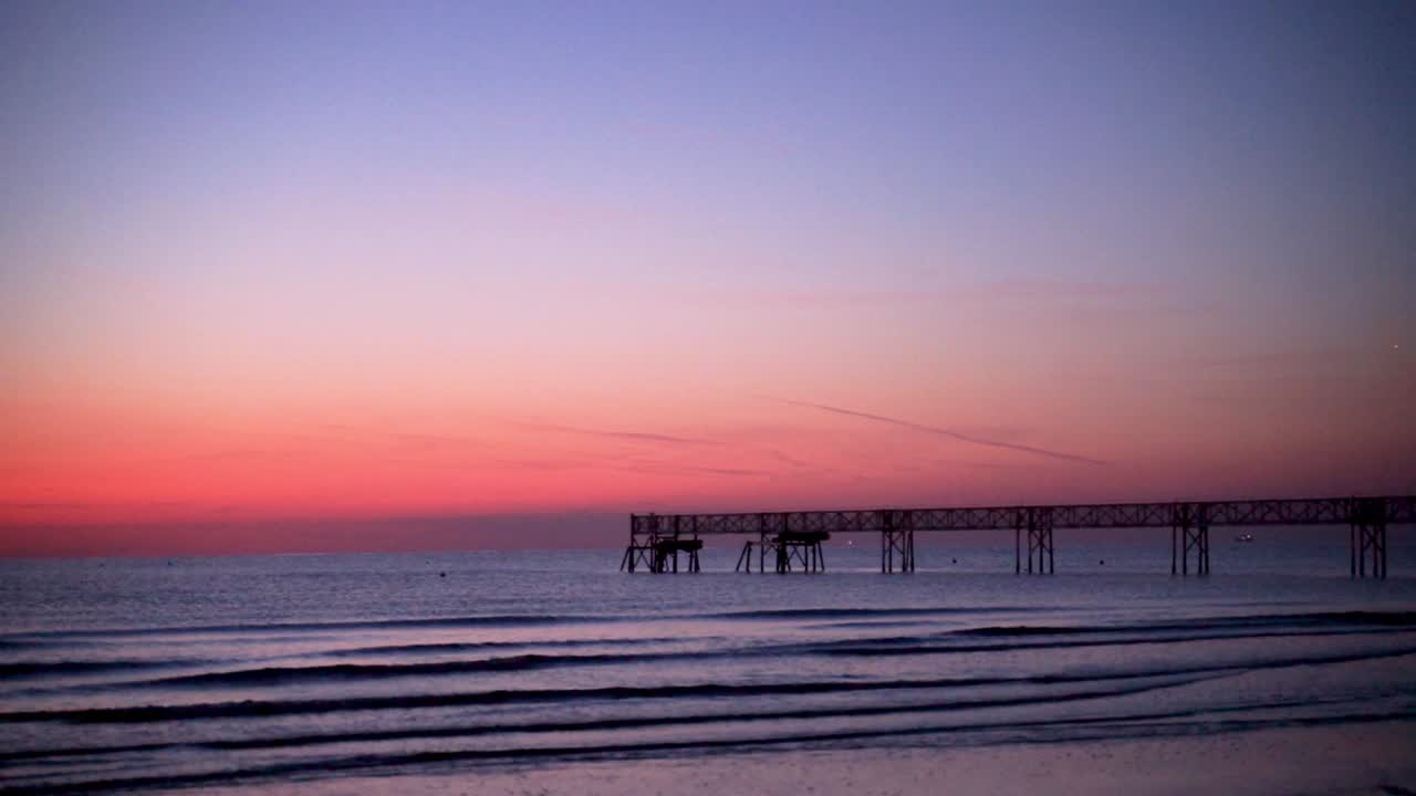 Dawn over a calm but sligthly wavy sea with a long pier in the middle of the scene