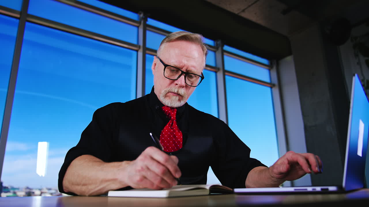 Serious mature businessman focused on work. Man types on laptop and writes in his paper notebook. Low angle view on the busy entrepreneur.