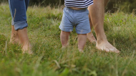 close up of a little baby taking his first steps while walking barefoot on the green grass and holding his parents' hands