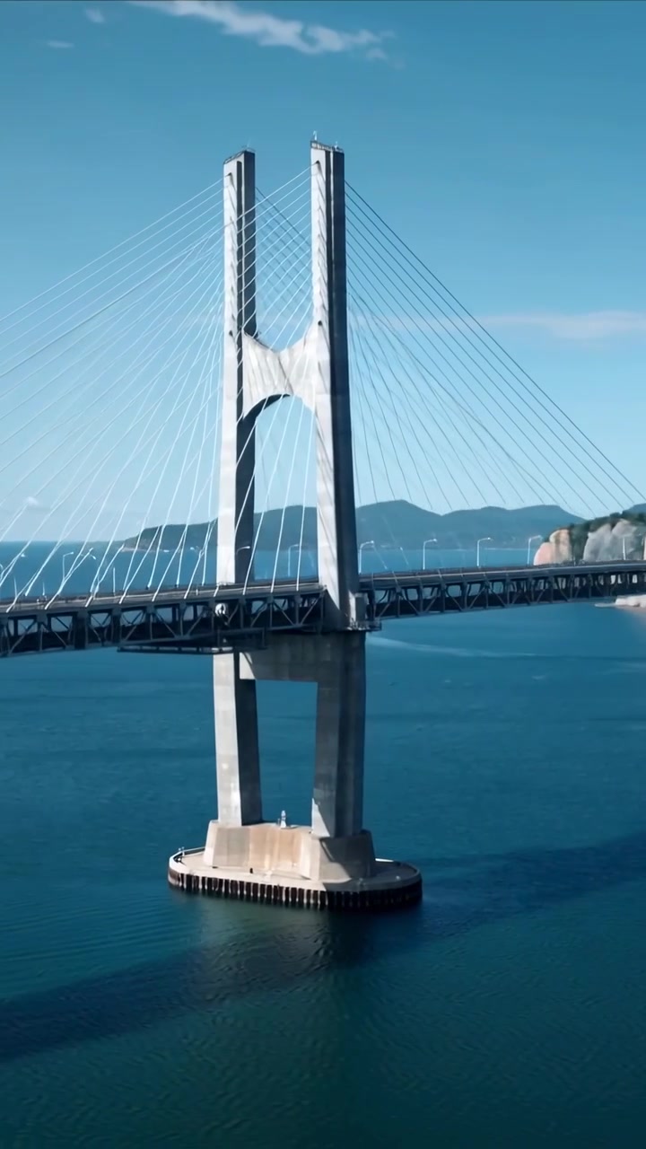 Video verticale: Drone che inizia un passaggio in avanti sopra una baia calma, rivelando la forma della torre di un ponte strallato