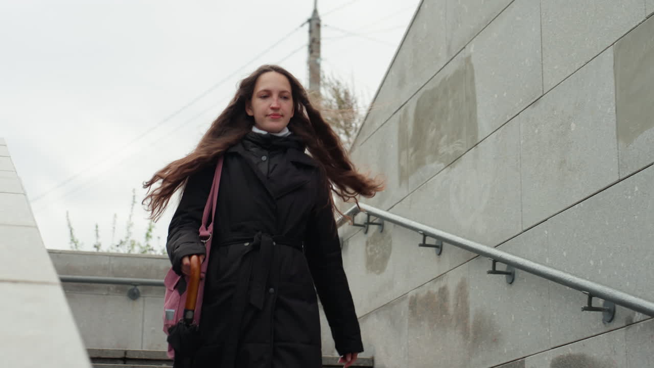 Donna che scende le scale di cemento con un ombrello. Pendolare urbana che scende sotto un cielo nuvoloso, indossa un lungo cappotto scuro e una sciarpa, passo sicuro oltre il corrimano in metallo e il muro di pietra ruvida, strada spontanea