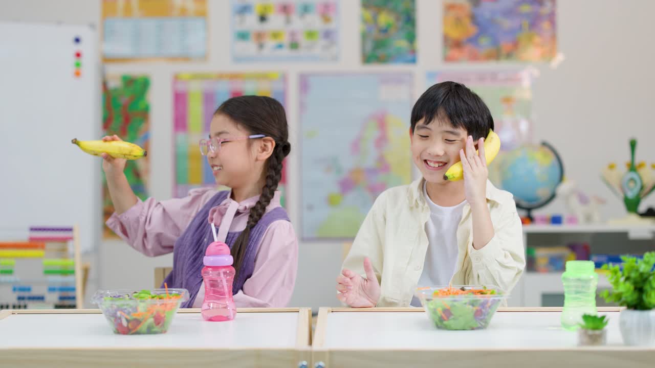 밝은 교실에서 샐러드를 먹으며 바나나 모양의 장난감 전화기로 장난치는 두 명의 아시아 아이들