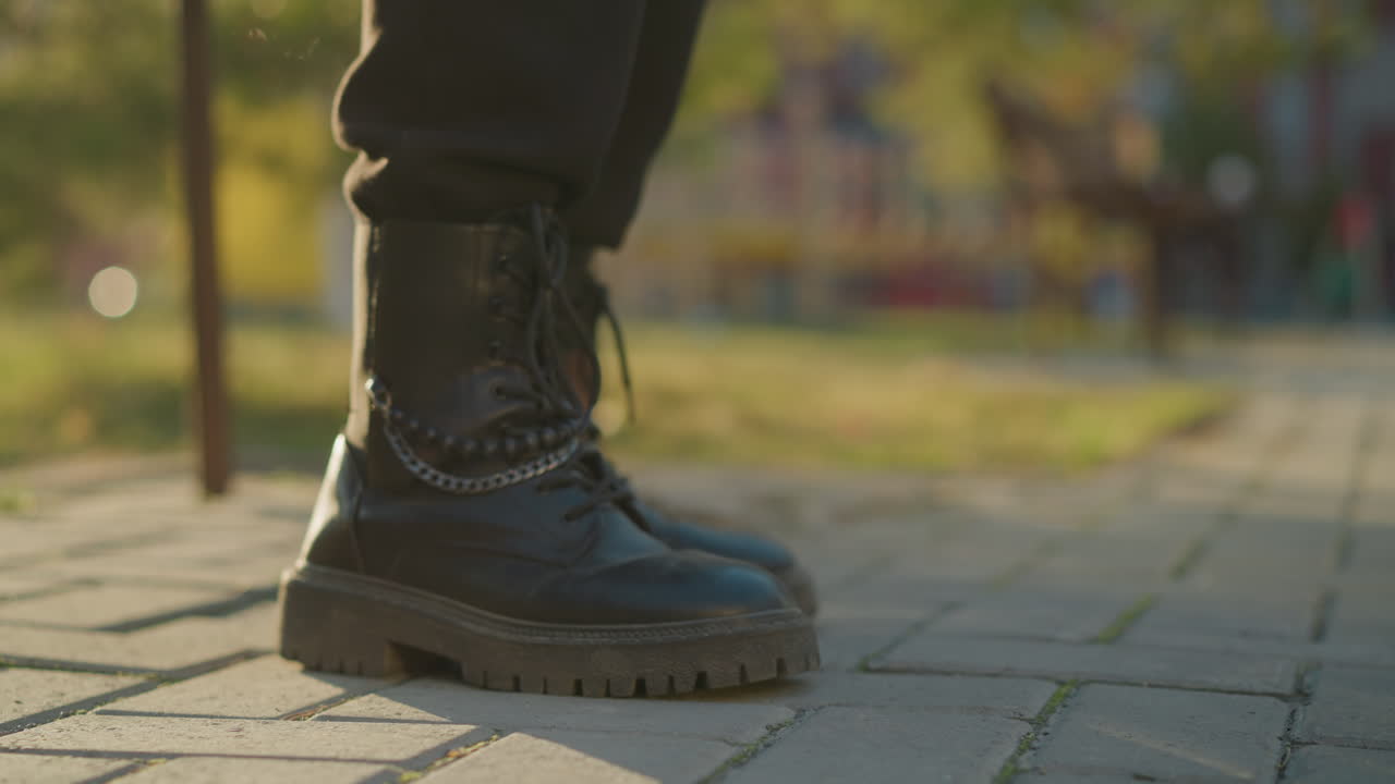 A close-up shot of black boots stepping on a sunlit paved path in a park. The focus is on one boot, with the background softly blurred, highlighting the outdoor setting with hints of greenery