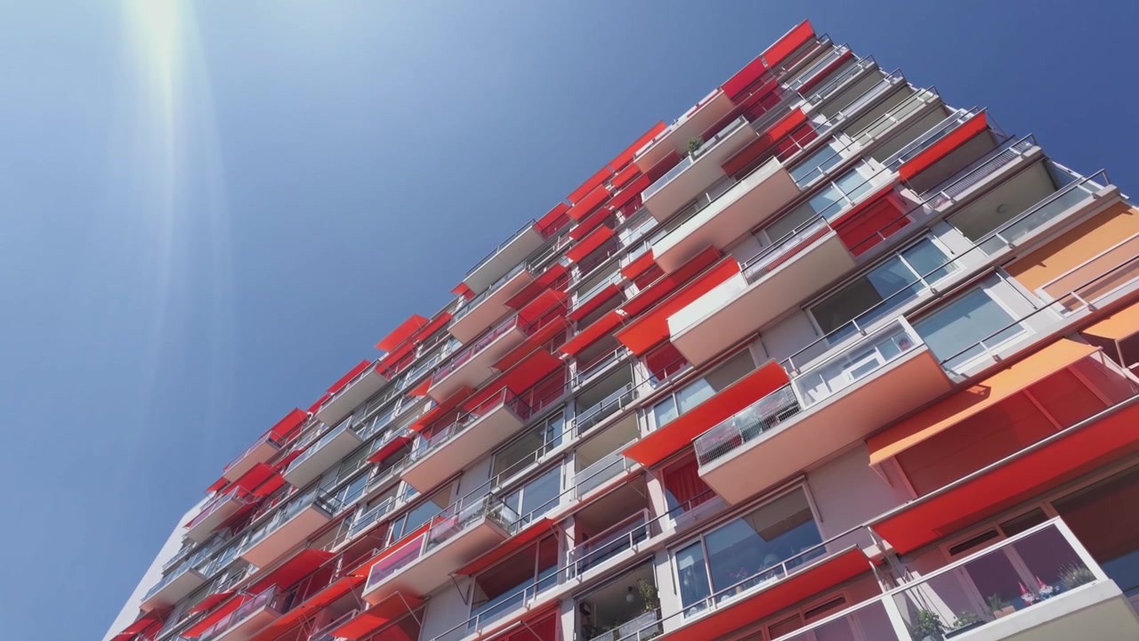 Plano aberto em ângulo baixo de um edifício residencial moderno de vários andares com muitas varandas e toldos vermelhos, a câmera inclina-se lentamente para cima. Um brilho solar intenso adiciona um toque atmosférico contra o céu azul claro.
