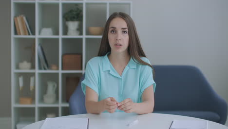 Head-shot-woman-sit-on-couch-look-at-camera-greeting-friend-start-video-call.-Young-adult-female-with-short-curly-hairstyle-enjoy-pleasant-conversation-by-video-conference-event-concept