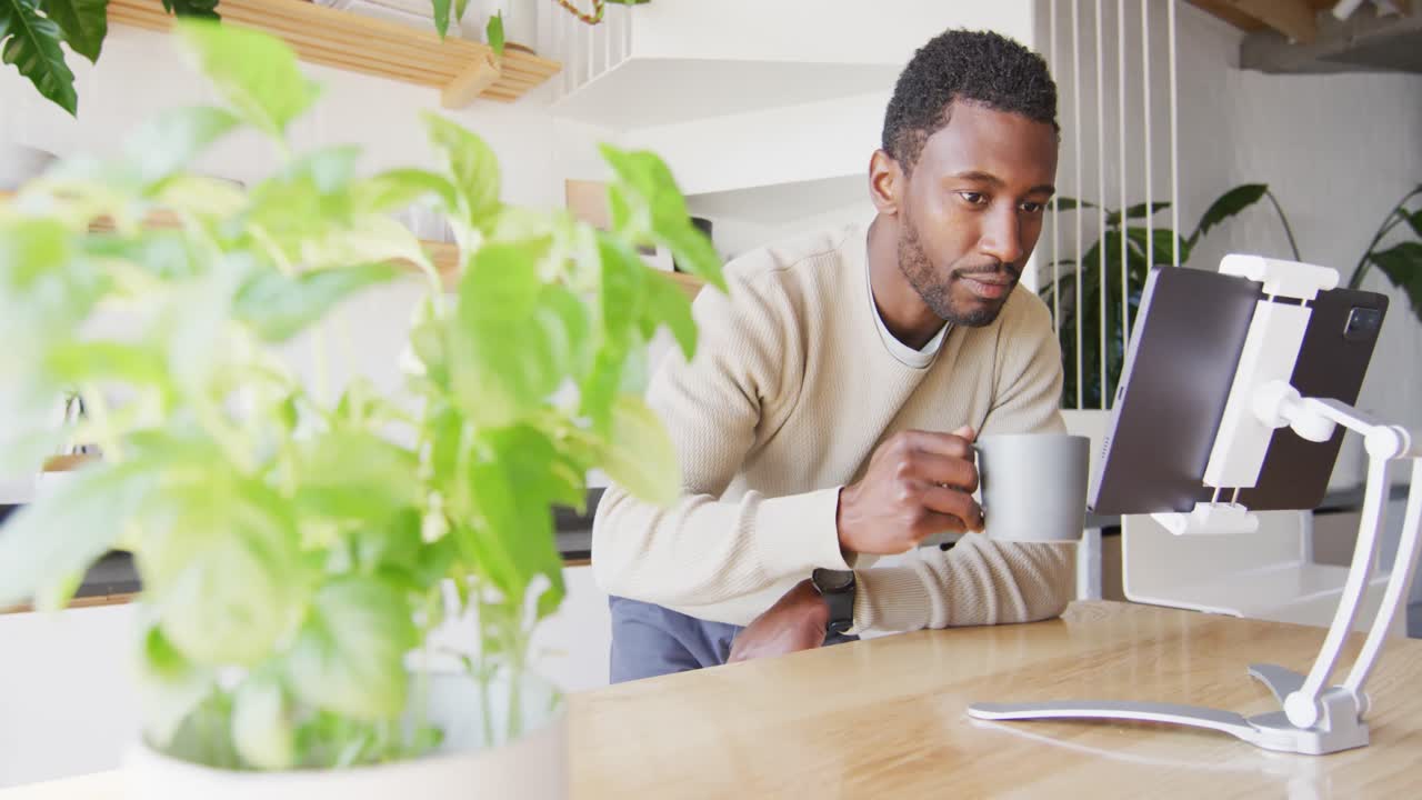 Happy african american man leaning on countertop in kitchen, using tablet and drinking coffee