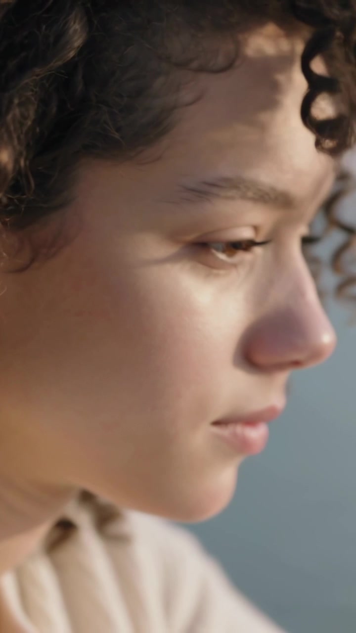 Vertical video: Breeze lifting curl, teen blinking and softening smile on shore, wearing hoodie