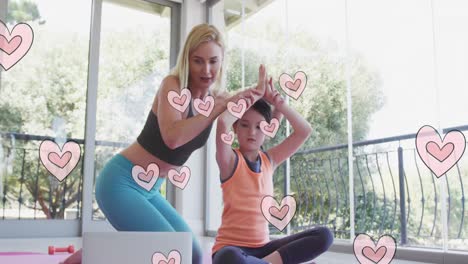 Animation-of-hearts-moving-over-happy-caucasian-woman-with-her-daughter-doing-yoga-using-laptop
