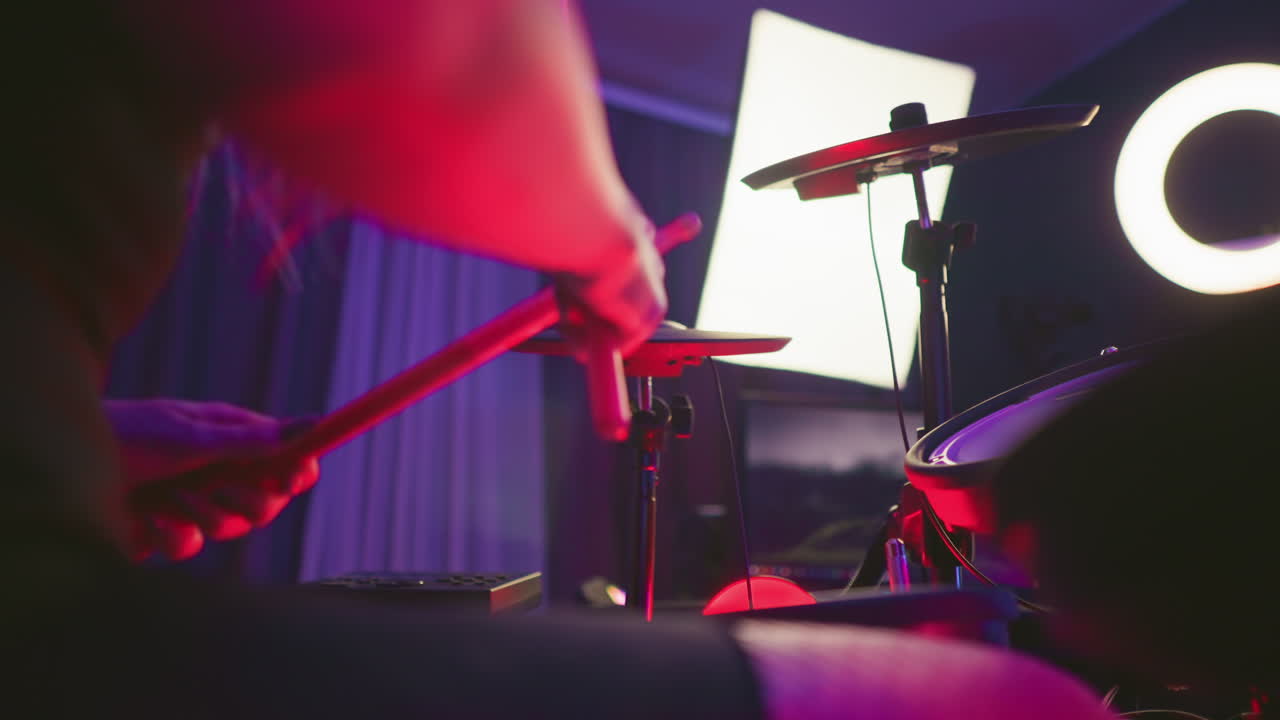 Close up of lady playing electronic drum set, striking cymbal while pressing pedal, under moody red and purple lighting with drum pads, monitor screen, and ring light visible in indoor studio setup